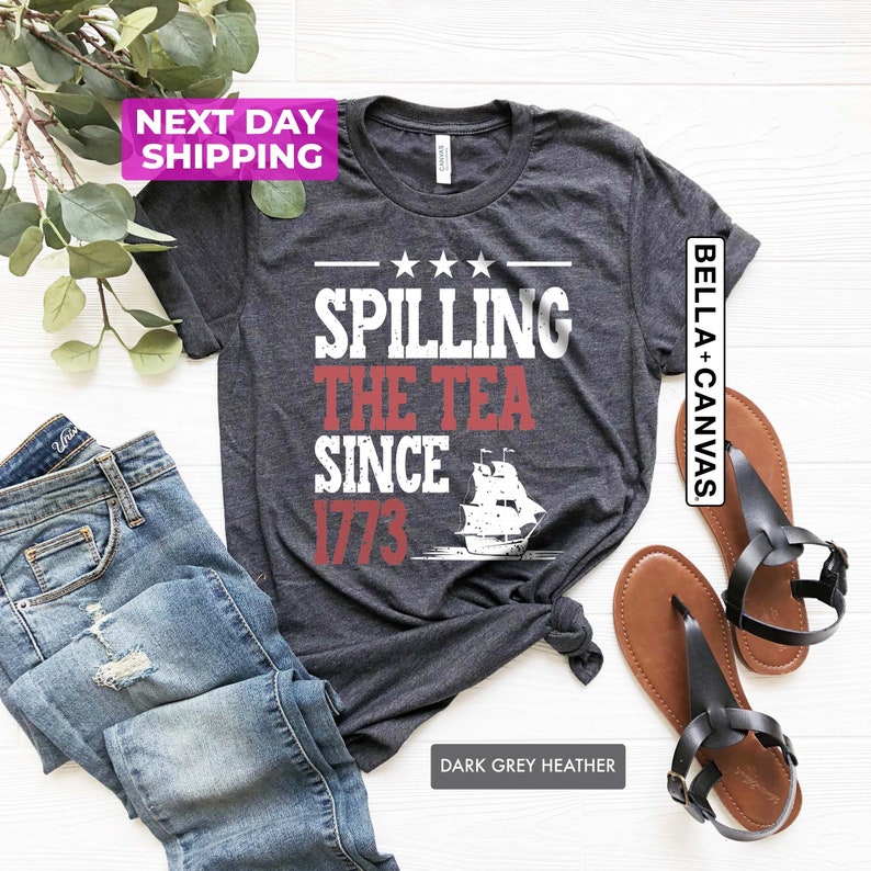 Pu&ograve; includere: Maglietta color grigio scuro melange con il testo "Spilling the Tea Since 1773" in rosso e bianco. Una silhouette bianca di una nave si trova sotto il testo. La maglietta &egrave; stesa su una superficie bianca.