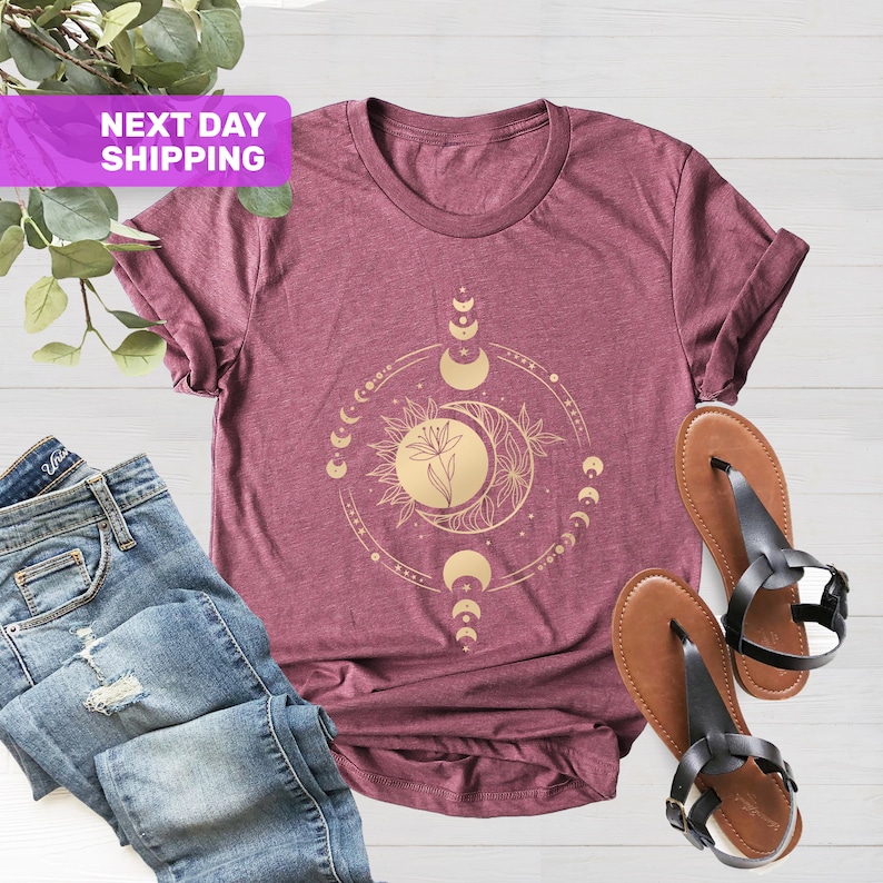 May include: A maroon t-shirt with a gold moon and sun graphic. The graphic features a flower in the center of the sun and moon. The moon phases are depicted around the sun and moon.