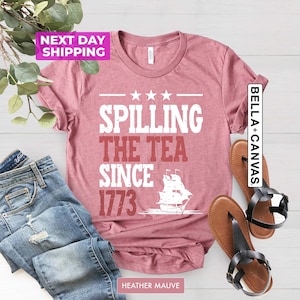 Pu&ograve; includere: Una t-shirt color malva con la scritta "Spilling the Tea Since 1773" e una silhouette bianca di una nave. La t-shirt &egrave; mostrata su una superficie bianca con un paio di sandali marroni e un paio di jeans blu.