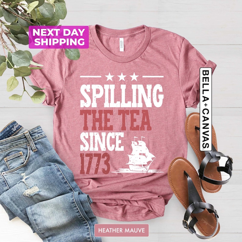 Pu&ograve; includere: Una t-shirt color malva con la scritta "Spilling the Tea Since 1773" e una silhouette bianca di una nave. La t-shirt &egrave; mostrata su una superficie bianca con un paio di sandali marroni e un paio di jeans blu.