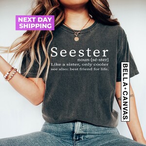 Pu&ograve; includere: Una maglietta grigia scura con la parola "Seester" definita come "Come una sorella, solo pi&ugrave; cool" e "vedi anche: migliore amico per la vita." La maglietta ha un logo bianco Bella + Canvas sul lato destro.