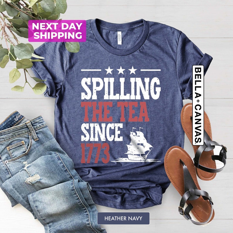 Pu&ograve; includere: Una maglietta blu navy con il testo "Spilling the Tea Since 1773" e una silhouette bianca di una nave. La maglietta &egrave; su una superficie bianca con un paio di sandali marroni e un paio di jeans blu.