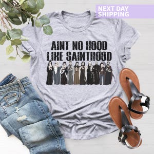K&ouml;nnte beinhalten: Graues T-Shirt mit dem Text "Ain't no hood like Sainthood" in schwarzer Schrift. Der Text ist von einer Reihe von Comicfiguren in Schwarzwei&szlig; umgeben.