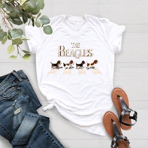 K&ouml;nnte beinhalten: Wei&szlig;es T-Shirt mit einer Grafik von f&uuml;nf Beagle-Hunden, die &uuml;ber einen Zebrastreifen laufen, mit dem Text "THE BEAGLES" dar&uuml;ber.