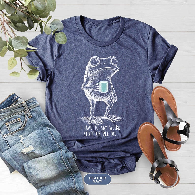 May include: Heather navy blue t-shirt with a white illustration of a frog holding a mug. The text "I HAVE TO SAY WEIRD STUFF OR I'LL DIE" is printed below the frog. The shirt is paired with blue jeans and brown sandals.