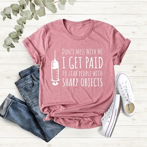 May include: A pink t-shirt featuring the text "DON'T MESS WITH ME I GET PAID TO STAB PEOPLE WITH SHARP OBJECTS" and a syringe illustration. The shirt is styled with blue jeans and white trainers.