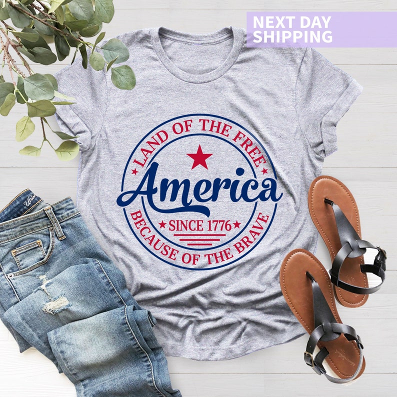 May include: Gray t-shirt with a red, white, and blue circular graphic that says "Land of the Free, America, Because of the Brave, Since 1776".