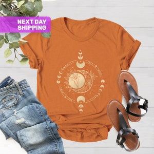 May include: A rust-colored t-shirt with a gold moon and sun graphic. The graphic features a flower and a crescent moon with a circle of smaller moons around it.