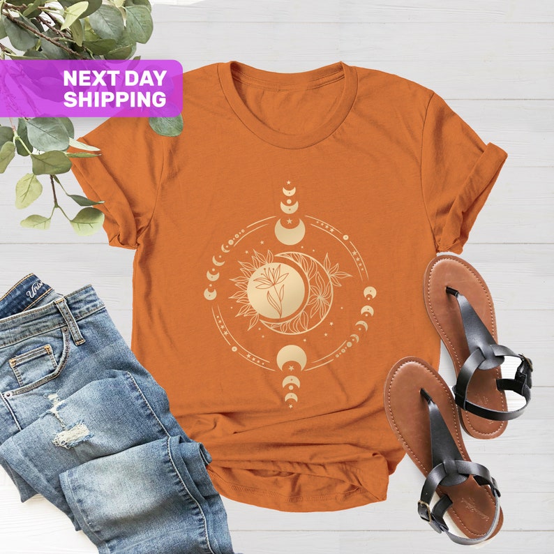 May include: A rust-colored t-shirt with a gold moon and sun graphic. The graphic features a flower and a crescent moon with a circle of smaller moons around it.