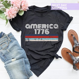 May include: Black t-shirt with a vintage-style graphic that says "AMERICA 1776" in white letters with red, white, and blue stripes underneath.