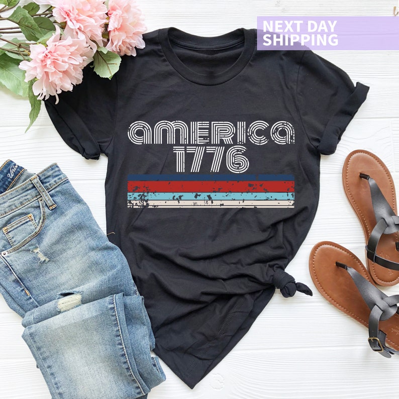 May include: Black t-shirt with a vintage-style graphic that says "AMERICA 1776" in white letters with red, white, and blue stripes underneath.