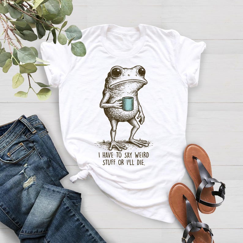 May include: White t-shirt featuring a detailed illustration of a frog holding a teal mug. The frog is drawn in a vintage style, with the text "I HAVE TO SAY WEIRD STUFF OR I'LL DIE" printed below. The shirt is paired with blue jeans and sandals.