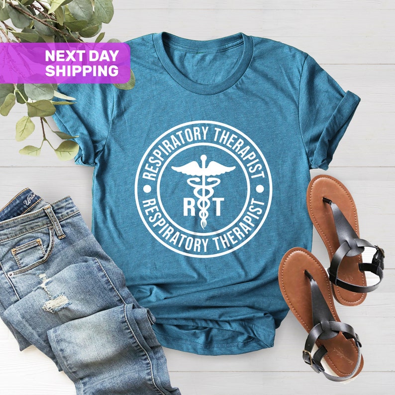 May include: Teal blue t-shirt with a white circle graphic featuring a medical symbol, the letters "RT", and the text "Respiratory Therapist".