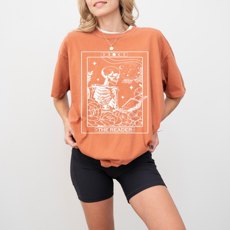 May include: A burnt orange t-shirt with a white graphic of a skeleton reading a book. The graphic is surrounded by flowers and stars. The text "THE READER" is below the graphic.
