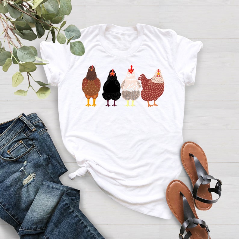 Peut inclure: T-shirt blanc avec un motif de quatre poulets en rang. Les poulets sont bruns, noirs, blancs et bruns avec des cr&ecirc;tes rouges.