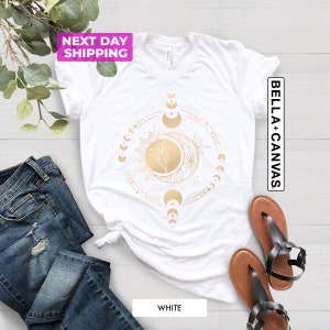 May include: White t-shirt with a gold moon and sun design. The design features a crescent moon with a star, a sun with a flower, and a circle of moons in different phases. The text "Bella + Canvas" is printed on the right side of the shirt.
