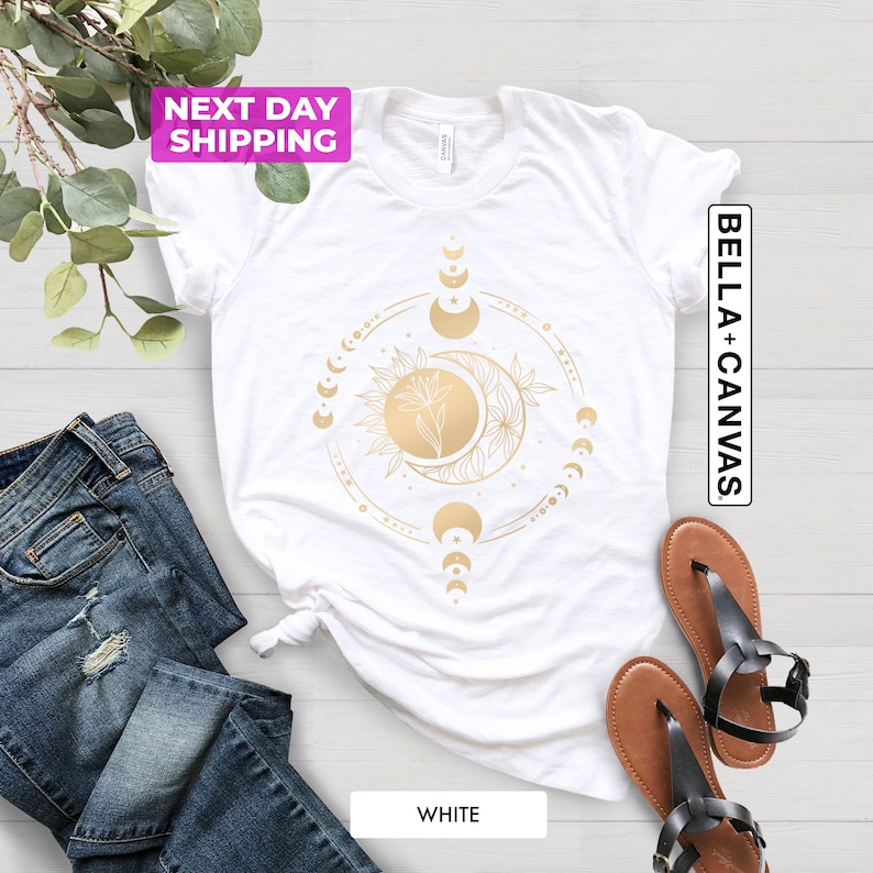 May include: White t-shirt with a gold moon and sun design. The design features a crescent moon with a star, a sun with a flower, and a circle of moons in different phases. The text "Bella + Canvas" is printed on the right side of the shirt.