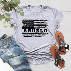May include: Gray t-shirt with a distressed American flag design and the text "Abuelo" in black. Below the flag are the names "Alice, Nicole, Sophia, Sarah, Daniel, Rebecca".