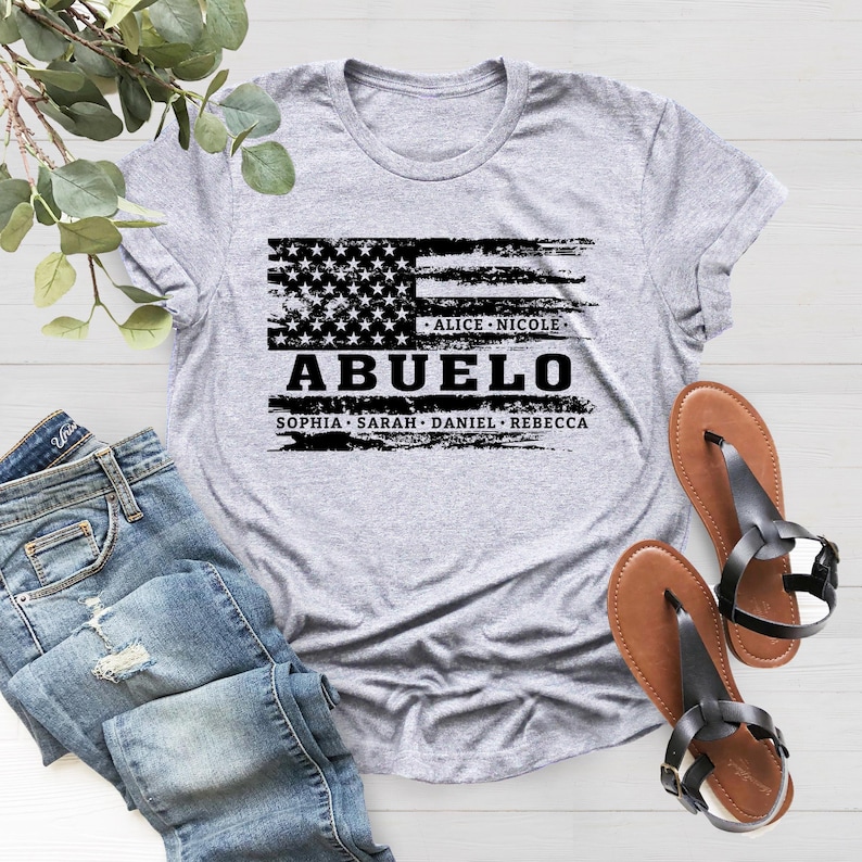 May include: Gray t-shirt with a distressed American flag design and the text "Abuelo" in black. Below the flag are the names "Alice, Nicole, Sophia, Sarah, Daniel, Rebecca".