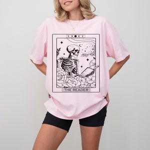 May include: Pink oversized t-shirt with a black and white tarot card design featuring a skeleton reading a book. The card reads "THE READER".