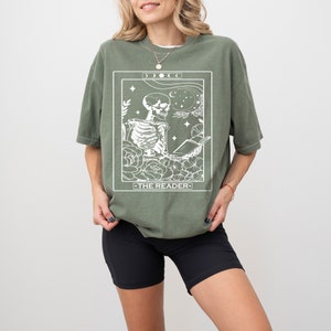 May include: A green t-shirt with a white graphic of a skeleton reading a book. The graphic is surrounded by flowers and stars. The text "THE READER" is below the skeleton.