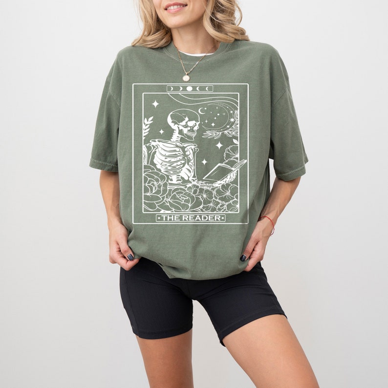 May include: A green t-shirt with a white graphic of a skeleton reading a book. The graphic is surrounded by flowers and stars. The text "THE READER" is below the skeleton.