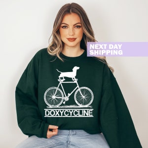May include: A dark green sweatshirt with a white graphic of a dachshund riding a bicycle. The text "DOXYCYCLINE" is printed below the bicycle.