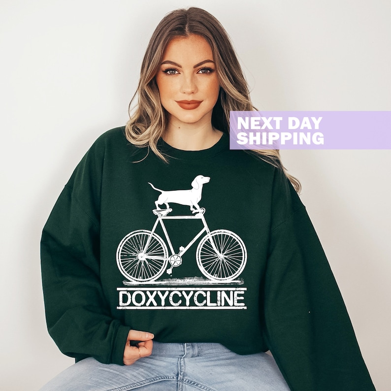 May include: A dark green sweatshirt with a white graphic of a dachshund riding a bicycle. The text "DOXYCYCLINE" is printed below the bicycle.