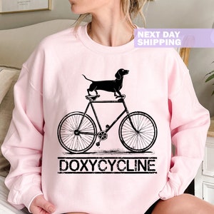 May include: A pink sweatshirt with a black dachshund riding a bicycle and the text "DOXYCYCLINE" underneath.