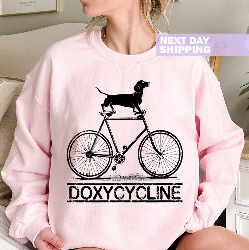 May include: A pink sweatshirt with a black dachshund riding a bicycle and the text "DOXYCYCLINE" underneath.