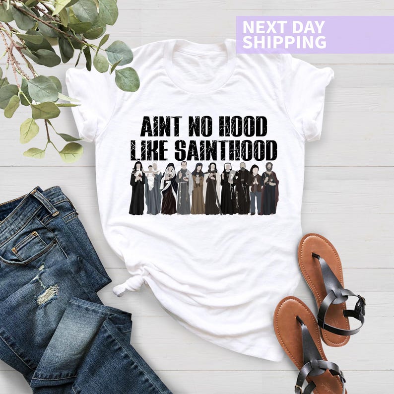 K&ouml;nnte beinhalten: Wei&szlig;es T-Shirt mit schwarzem Text "Ain't no hood like Sainthood" und einer Grafik einer Gruppe von Menschen in religi&ouml;ser Kleidung.