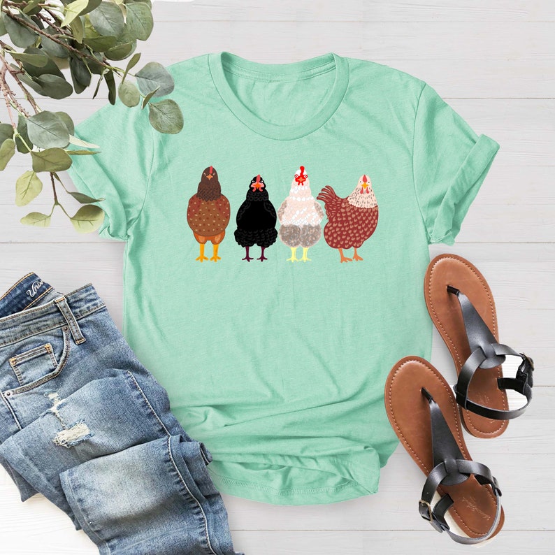 Peut inclure: Un t-shirt vert clair avec un motif de quatre poulets en rang. Les poulets sont bruns, noirs, blancs et rouges.