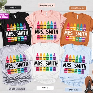 May include: A set of ten t-shirts in various colors featuring a rainbow of crayons with the text "MRS. SMITH" printed on each shirt.