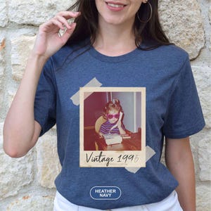 May include: Heather navy t-shirt with a vintage photo print. The photo shows a young girl wearing heart-shaped sunglasses and talking on a telephone. The words "Vintage 1995" are printed below the photo. The shirt also has the words "Heather Navy" printed on it.