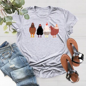 Peut inclure: Un t-shirt gris avec un motif de quatre poulets en rang. Les poulets sont bruns, noirs, blancs et rouges.