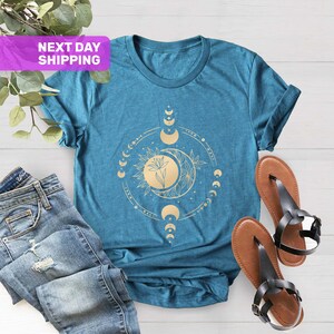 May include: Teal blue t-shirt with a gold moon and flower design. The design features a circle of moons in different phases, with a flower in the center.