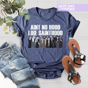 K&ouml;nnte beinhalten: Ein marineblaues T-Shirt mit dem Text "Ain't no hood like Sainthood" in wei&szlig;en Buchstaben. Das Shirt zeigt eine Grafik einer Gruppe von Menschen in religi&ouml;ser Kleidung.