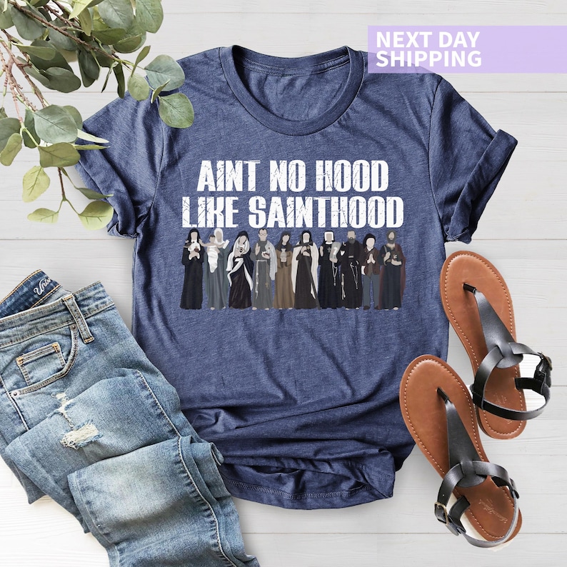 K&ouml;nnte beinhalten: Ein marineblaues T-Shirt mit dem Text "Ain't no hood like Sainthood" in wei&szlig;en Buchstaben. Das Shirt zeigt eine Grafik einer Gruppe von Menschen in religi&ouml;ser Kleidung.