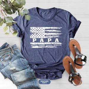 May include: A navy blue t-shirt with a distressed white American flag design. The text "PAPA" is printed below the flag, with the names "Alice, Nicole, Sophia, Sarah, Daniel, Rebecca" printed below the text. The shirt is available in heather navy.