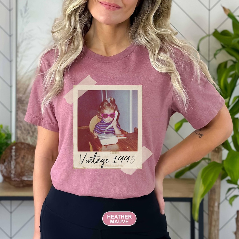 May include: Heather mauve t-shirt featuring a vintage photo print. The photo shows a child wearing sunglasses and using a telephone. The text "Vintage 1995" is below the photo. The shirt is a solid color.
