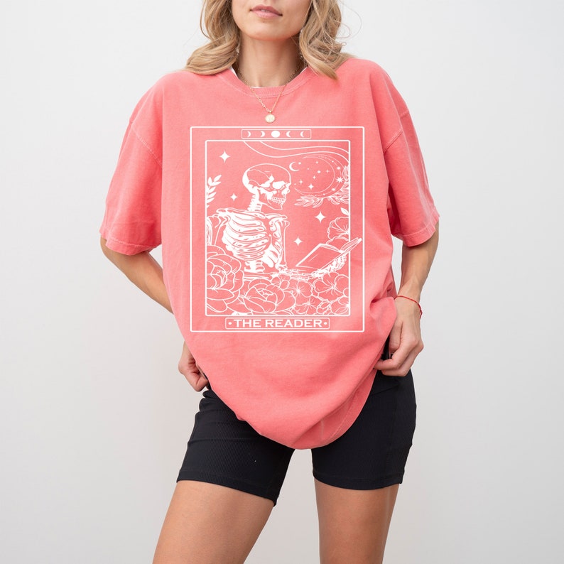 May include: A coral pink oversized t-shirt with a white graphic of a skeleton reading a book in a floral setting. The graphic is framed with a white border and includes the text "THE READER".