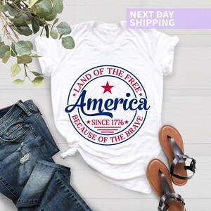May include: White t-shirt with a red, white, and blue circular graphic that says "Land of the Free, America, Since 1776, Because of the Brave".
