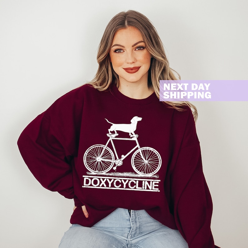 May include: A maroon sweatshirt with a white graphic of a dachshund riding a bicycle. The text "DOXYCYCLINE" is printed below the graphic.