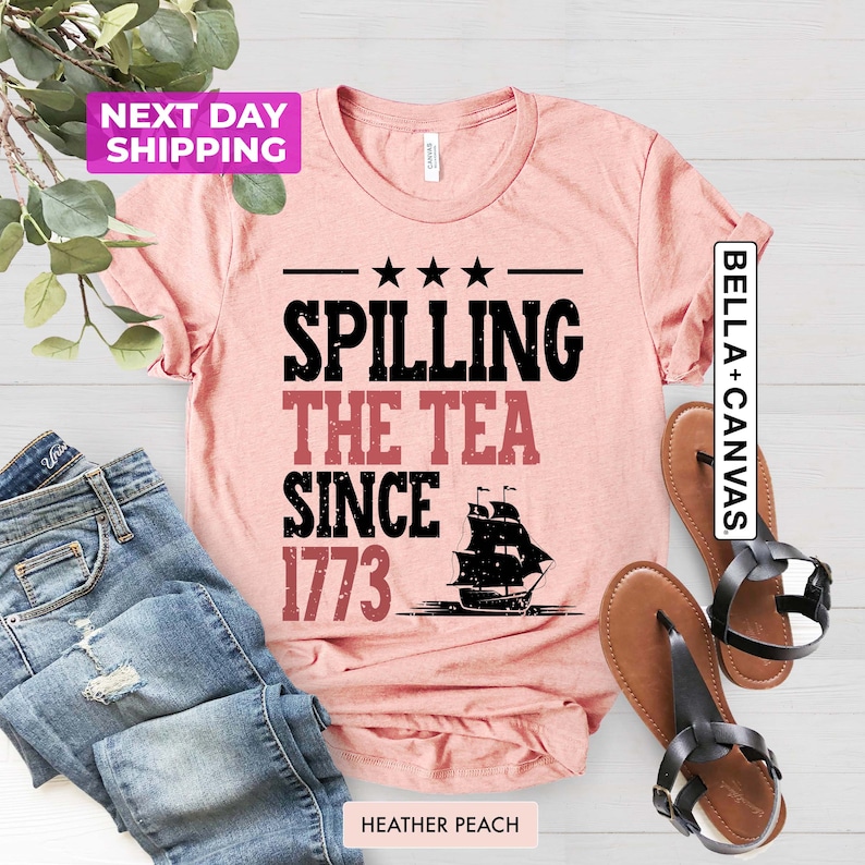 Pu&ograve; includere: Una maglietta rosa con il testo "Spilling the Tea Since 1773" in nero con una silhouette nera di una nave. La maglietta &egrave; su una superficie di legno con un paio di sandali marroni.