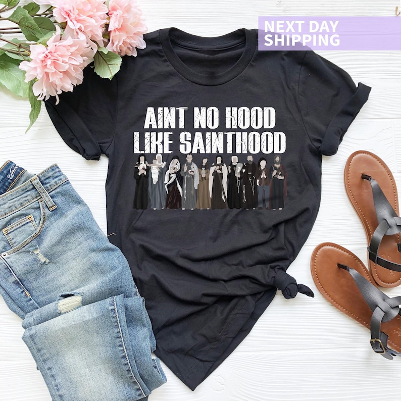 K&ouml;nnte beinhalten: Schwarzes T-Shirt mit einer wei&szlig;en Grafik, auf der "Ain't no hood like Sainthood" steht und eine Reihe von Comicfiguren in Schwarzwei&szlig; abgebildet sind.
