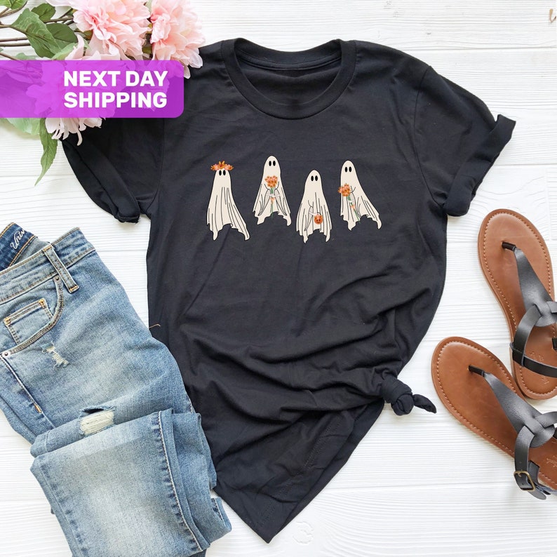以下が含まれることがあります： 4体の幽霊が花を持っているグラフィックがプリントされた黒いTシャツ。幽霊は白で、目は黒。花はオレンジと黄色です。