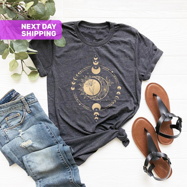 May include: A dark grey t-shirt with a gold floral moon phase design. The design features a stylized flower with a crescent moon and sun.