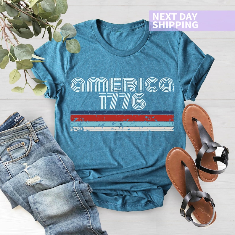 May include: A teal blue t-shirt with a white graphic that says "America 1776" with red, white, and blue stripes underneath.