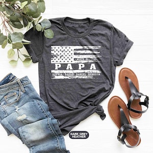 May include: Dark grey heather t-shirt with a distressed American flag design. The text "PAPA" is printed in white with the names "Alice, Nicole, Sophia, Sarah, Daniel, Rebecca" printed below.