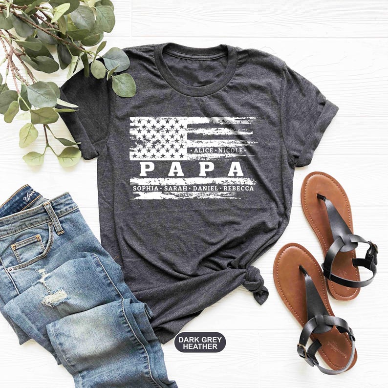 May include: Dark grey heather t-shirt with a distressed American flag design. The text "PAPA" is printed in white with the names "Alice, Nicole, Sophia, Sarah, Daniel, Rebecca" printed below.